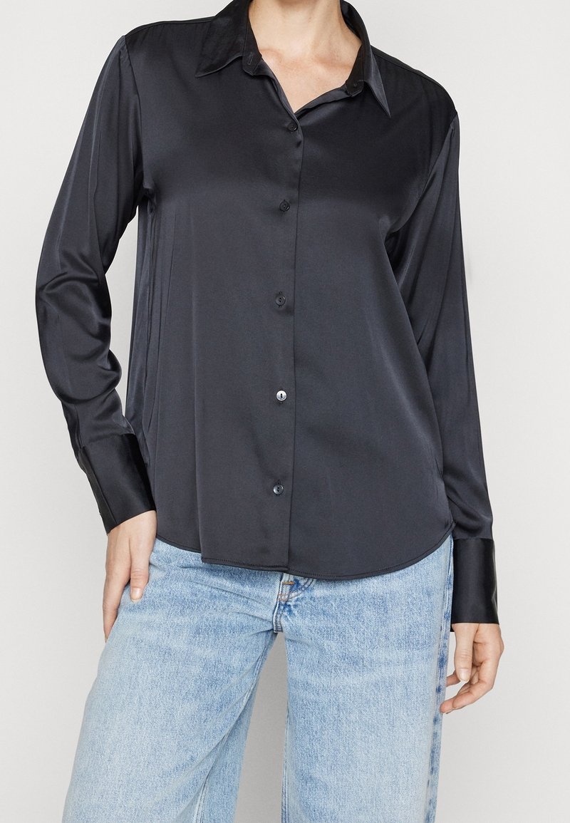 Zwarte satijnen langärmelige blouse met een knoopsluiting aan de voorkant, een klassieke kraag en een soepele textuur. Gecombineerd met lichtblauwe denim jeans.