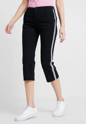 Femme portant un pantalon noir court avec des bandes blanches sur les côtés, un haut rose et des baskets blanches, debout sur un fond uni.