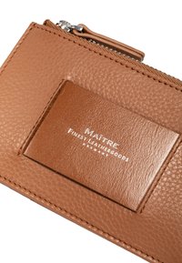 Portafoglio in pelle marrone con superficie texturizzata, dotato di una patchetta con logo in rilievo argentato e chiusura a zip lungo il bordo superiore.
