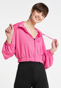 Rosa cropped zip-up jakke lavet af mesh stof, med elastiske manchetter og en indsnævret talje, parret med sorte leggings.