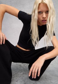 Camiseta negra ajustada y pantalones deportivos sueltos negros en un modelo en posición de sentadilla baja. Cabello rubio liso y ondulado con un fondo neutro.