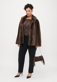 Brązowy płaszcz z futra sztucznego z szerokim kołnierzem, brązowa bluzka, czarne spodnie, czarne szpilki oraz mała ciemna torebka z frędzlami.