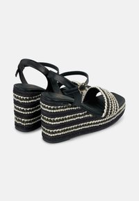 Zwarte wedge sandalen met witte en beige gevlochten details, voorzien van een gestructureerde geweven hak en verstelbare enkelbandjes.