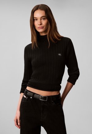 Kvinde med langt brunt hår iført en sort ribbet rullekravesweater og sorte bukser med sort bælte mod en neutral baggrund.