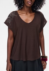 Brązowa, krótka koszulka z dekoltem w serek, wykonana z miękkiego materiału, ozdobiona koralikami na jednym ramieniu. Luźny krój.