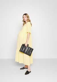 Vestido de maternidade amarelo fluido com mangas curtas bufantes, barra em camadas e um corte descontraído. Um clutch estruturado preto e sapatos slip-on pretos completam o visual.