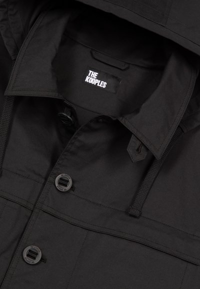 Schwarze, leichte Jacke aus glattem Stoff mit Kapuze, Knopfverschluss und einem Etikett mit der Aufschrift "The Kooples" innen.