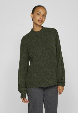 Pull en laine vert foncé avec un col montant, des poignets côtelés et une coupe loose, associé à un jean gris. Texture douce et silhouette décontractée.