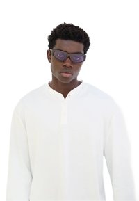 Camicia bianca in cotone a maniche lunghe con scollatura henley e tre bottoni; abbinata a occhiali da sole oversize scuri con una leggera sfumatura viola.