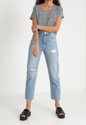 Kobieta nosząca szarą kraciastą bluzkę z krótkim rękawem, jasnoniebieskie spodnie jeansowe z wysokim stanem i efektami przetarć oraz czarne sandały na platformie, stoi ze skrzyżowanymi ramionami.