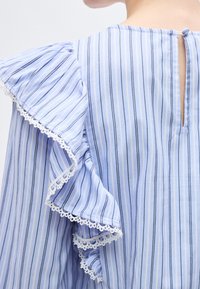 Μπλούζα με κάθετες μπλε και λευκές ρίγες, μανίκια με βολάν και τελείωμα από λευκή δαντέλα, με κλειδί στο πίσω μέρος για κλείσιμο.