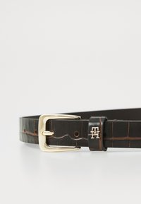 Ceinture en cuir noir avec une texture crocodile, boucle rectangulaire en or et un petit logo doré près de la boucle.
