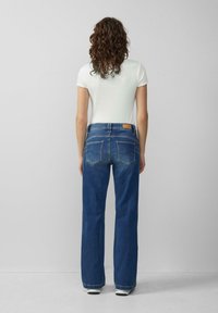 Frau mit lockigem Haar steht mit dem Rücken zur Kamera, trägt ein weißes T-Shirt mit kurzen Ärmeln, hochgeschnittene blaue Jeans und weiße Schuhe vor einem schlichten Hintergrund.