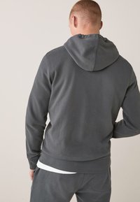 Sudadera gris con capucha, de textura suave, que cuenta con un cordón en la capucha y puños acanalados. Combinada con pantalones grises a juego y una camiseta blanca debajo.
