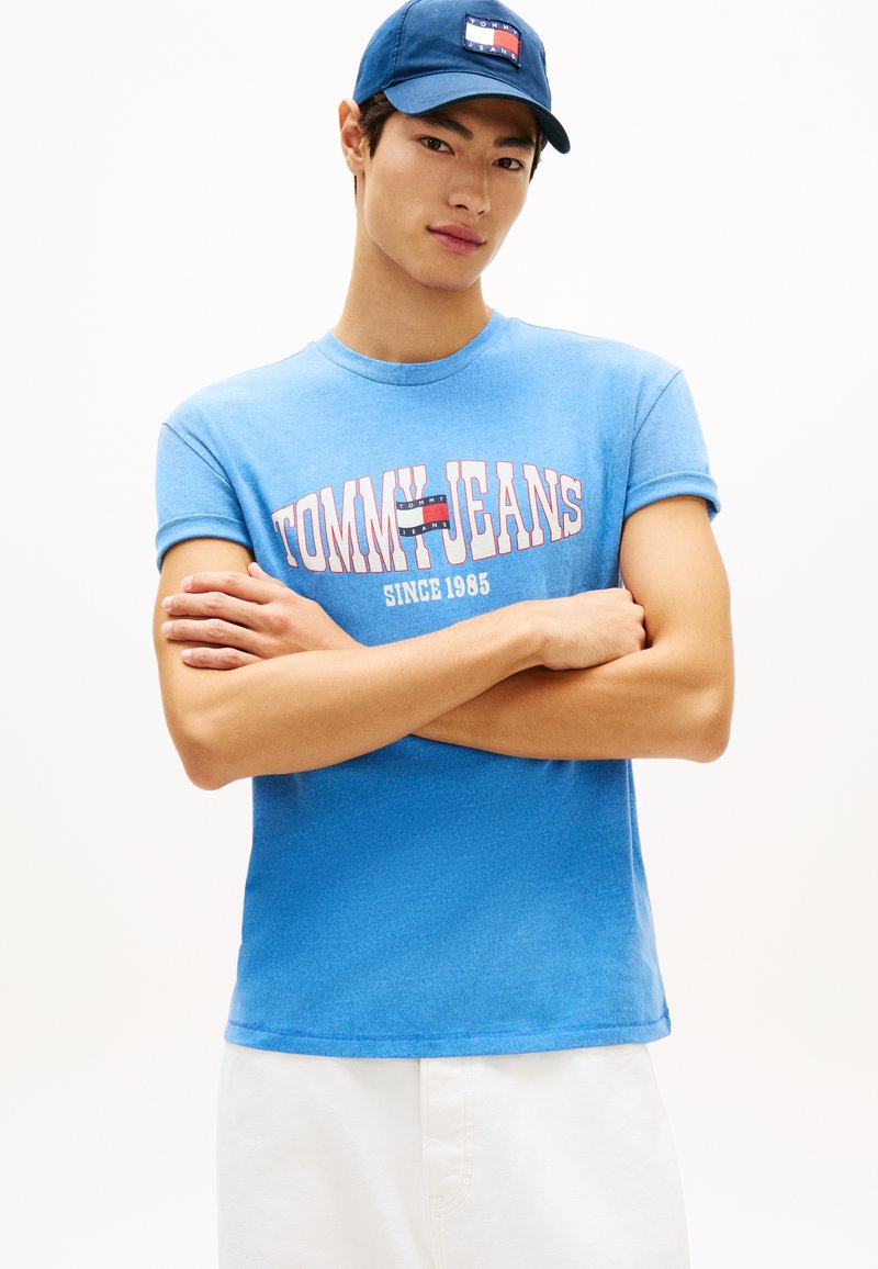 Νεαρός άνδρας που φοράει μπλε μπλουζάκι Tommy Jeans και σκούφο ναυτικού χρώματος, με σταυρωμένα χέρια, ποζάροντας μπροστά σε απλό λευκό φόντο.