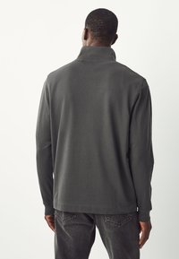 Maglia a maniche lunghe di colore grigio scuro con colletto alto, realizzata in tessuto morbido. Presenta una vestibilità comoda e cuciture flatlock. Indossata con denim scuro.