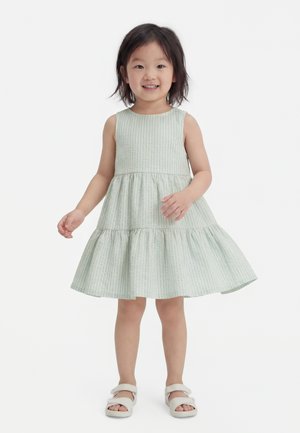 Giovane ragazza che sorride e gira in un vestito verde chiaro senza maniche con sandali su sfondo bianco.