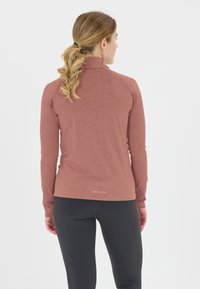 Kvindelig med blond hestehale iført en langærmet atletisk top i rosenfarve og sorte leggings, set bagfra mod en hvid baggrund.