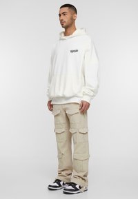 Sudadera blanca con capucha y un bolsillo frontal, combinada con pantalones cargo beige que tienen múltiples bolsillos. Zapatillas negras y blancas completan el look.