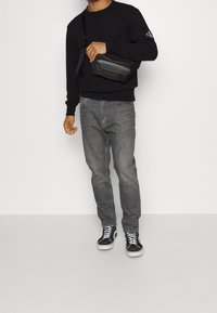 Felpa nera, jeans grigi e sneakers neri; l'uomo tiene una borsa a tracolla nera. L'outfit presenta un look casual, con una vestibilità rilassata e texture.
