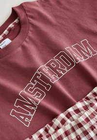 Bordeauxrode katoenen sweatshirt met de tekst "AMSTERDAM" in witte letters. Het onderste gedeelte heeft een rood-wit geruit patroon. Gestructureerde geribbelde halslijn.