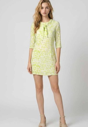 Joven mujer de pie vestida con un vestido verde claro con estampado, mangas tres cuartos y sandalias beige con tacón cuadrado.