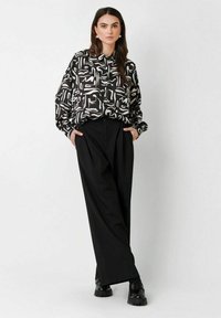 Woman with long dark hair in black and white abstract patterned blouse, wide black trousers, black chunky shoes, standing with hands in pockets.