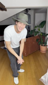 Šedý klobouk se širokým krempou a hnědým páskem, bílá tričko, šedé džíny a bílé tenisky. Vnitřní prostředí s hnědou pohovkou a rostlinou v květináči.