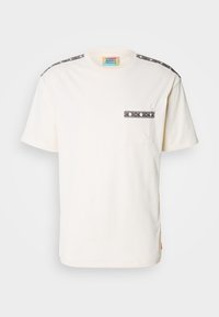 White cotton t-shirt with short sleeves, features a chest pocket and black patterned accents on the shoulders. Tag visible at the neckline.