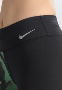 Zbliżenie na osobę w czarnych legginsach sportowych z szarym logo Nike na pasku i częściowym zielonym graficznym detalem.