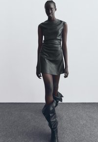 Rochie mini din piele neagră, cu o formă ajustată și o textură netedă. Cizme negre până la genunchi purtate lejer în jurul gleznelor. Fundal neutru.