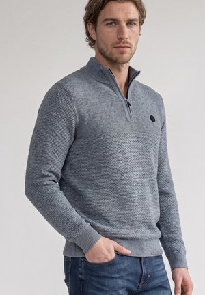 Homme portant un pull en maille gris texturé avec une fermeture éclair au quart et un jean bleu, debout devant un fond clair uni.