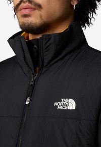 Chaqueta negra aislante con cuello alto, cremallera frontal y un logo blanco en el pecho. Interior forrado con material naranja.