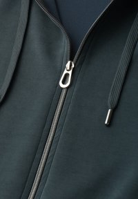 Sweat à capuche vert foncé avec fermeture éclair argentée partiellement fermée et cordons assortis avec embouts métalliques.
