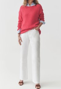 Korallenroter gestrickter Pullover mit lockerer Passform und hochgekrempelten Ärmeln, kombiniert mit weißen Hosen und braunen Sandalen mit Riemchen, vor einem schlichten Hintergrund.