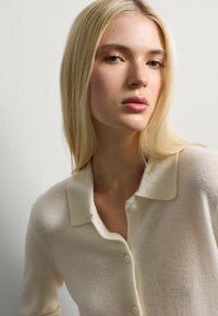 Donna bionda con capelli lisci che indossa un maglione crema con bottoni, che guarda leggermente di lato su uno sfondo bianco uniforme.