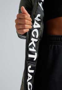 Olivengroen gestikt jasje met een rits aan de voorkant. Het binnenmateriaal heeft bold witte letters. Zwarte stof zichtbaar aan de onderkant.