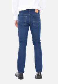 Uomo che indossa jeans blu, scarpe nere e una camicia bianca a maniche lunghe, in piedi con le mani vicino alle tasche, visto di spalle su uno sfondo bianco.