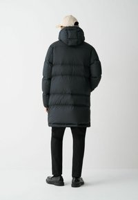 Črn puhovka z kapuco in prešitim vzorcem, s daljšim krojem, v kombinaciji s črnimi hlačami in črnimi čevlji, na enolični ozadju.