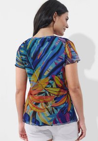Transparente Bluse mit lebendigem tropischen Blumenmuster in Blau, Gelb und Orange. Rundhalsausschnitt und kurze Ärmel mit gewellten Kanten.