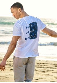White cotton t-shirt featuring a blue graphic design with the text "Edge of the Horizon" on the back, worn with light beige pants.