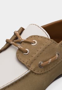 Primer plano de un zapato náutico de gamuza color canela y cuero blanco con cordones trenzados color canela pasados por ojales plateados en el panel lateral.
