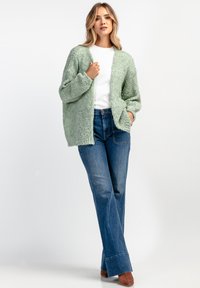 Jasnozielony, teksturowany, oversize'owy kardigan z długimi rękawami i otwartym przodem, noszony na białej bluzce i niebieskich dżinsach o wysokim stanie. Brązowe buty na block heel.