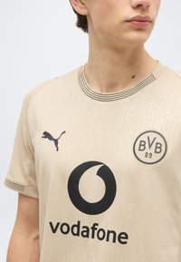 Νεαρός που φοράει ανοιχτό μπεζ ποδοσφαιρική φανέλα της Borussia Dortmund με λογότυπο Puma και χορηγό Vodafone στο στήθος.