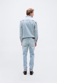 Φωτεινό μπλε τζιν μπουφάν και τζιν παντελόνι, χαρακτηρισμένα από κλασικό σχήμα, ραμμένες λεπτομέρειες και λεία υφή. Καφέ δερμάτινα παπούτσια ολοκληρώνουν την εμφάνιση.