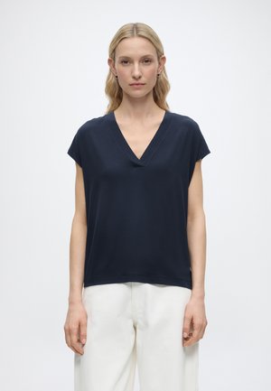 Mujer rubia con el pelo suelto, vestida con una camiseta azul marino de cuello en V y manga corta, y pantalones blancos, de pie frente a un fondo liso.