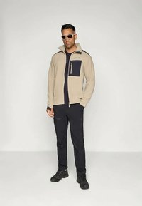 Light beige zip-up fleece jacket with navy pocket, paired with black pants and shoes. Model with sunglasses stands against a plain background.