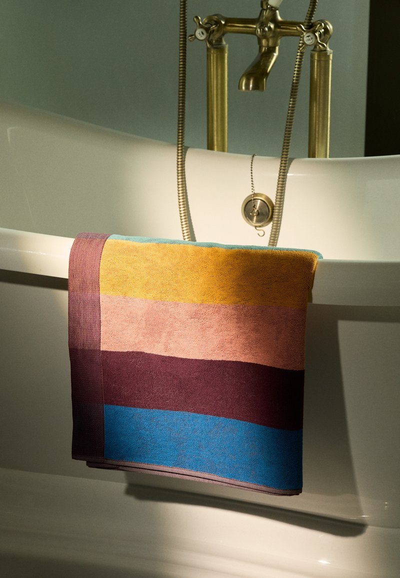 Serviette multicolore présentant des rayures horizontales dans des teintes de jaune, rose, violet et bleu, fabriquée en tissu texturé, drapée sur une baignoire.