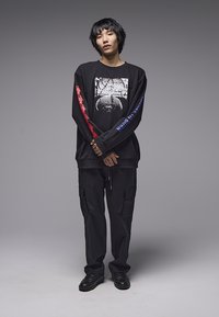 Μαύρο μακρυμάνικο πουκάμισο με γραφικό σχέδιο και κόκκινες και μπλε λεπτομέρειες· σετ με φαρδιά μαύρη παντελόνια καρό και μαύρα αθλητικά παπούτσια.