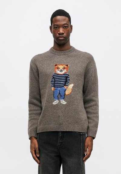 Vīrietis, kurš valkā pelēku džemperi ar pūci ar svītrainu krekliņu, džinsus un sporta apavus, ar adītu motīvu priekšā, kombinēts ar melniem džinsiem.